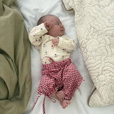Newborn baby with floral bodysuit and red/white gingham pants lying on a bed