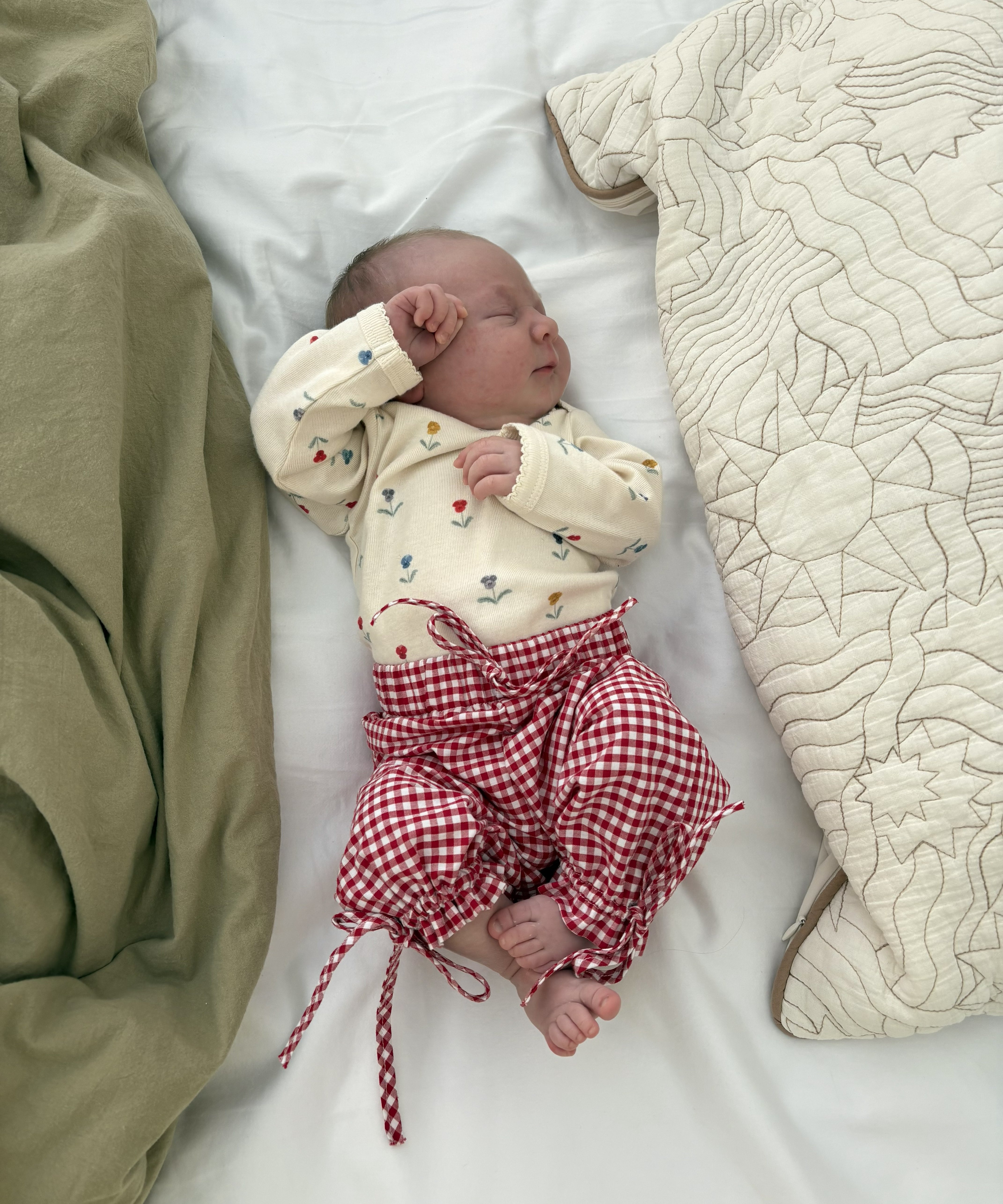 Newborn baby with floral bodysuit and red/white gingham pants lying on a bed