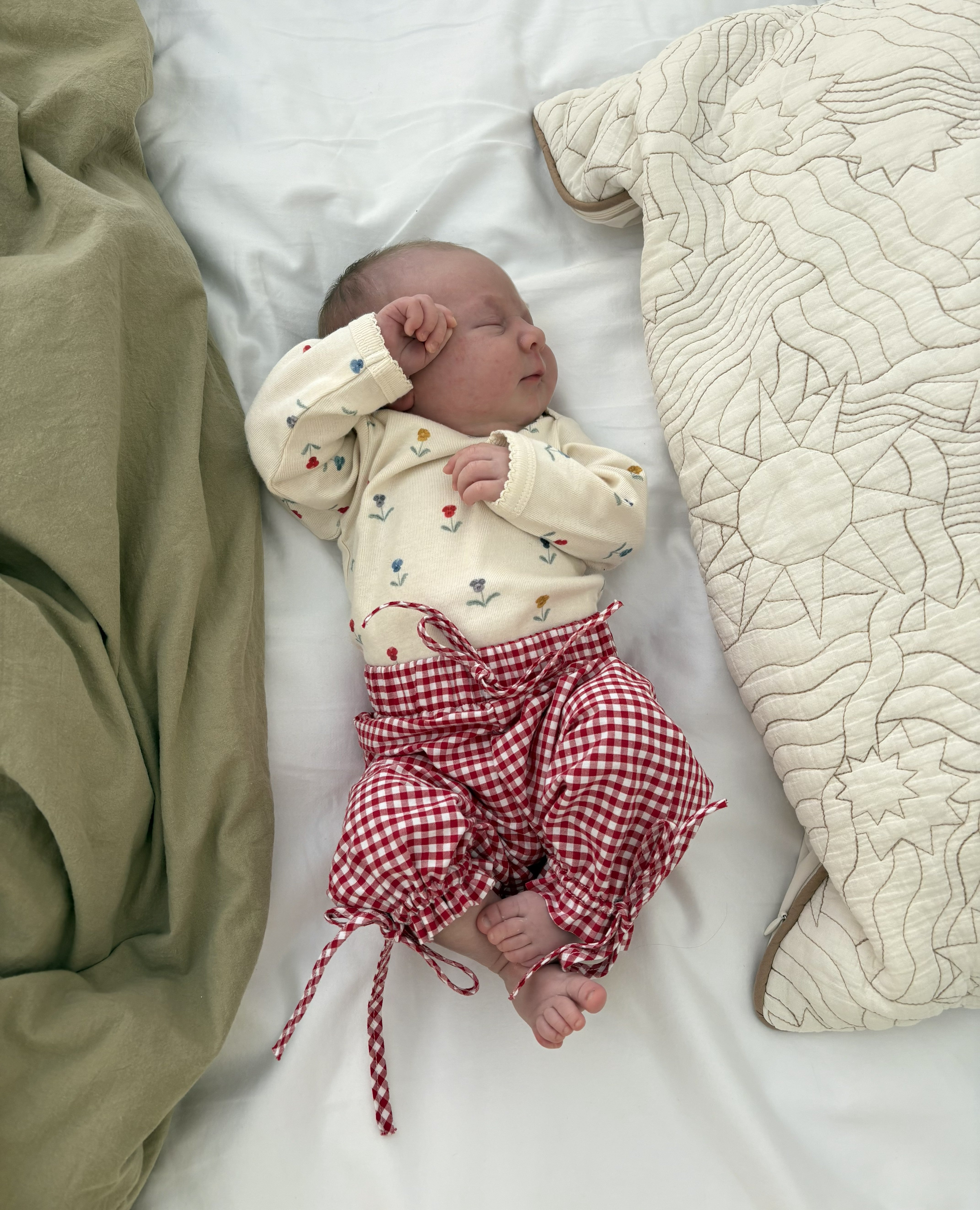 Newborn baby with floral bodysuit and red/white gingham pants lying on a bed