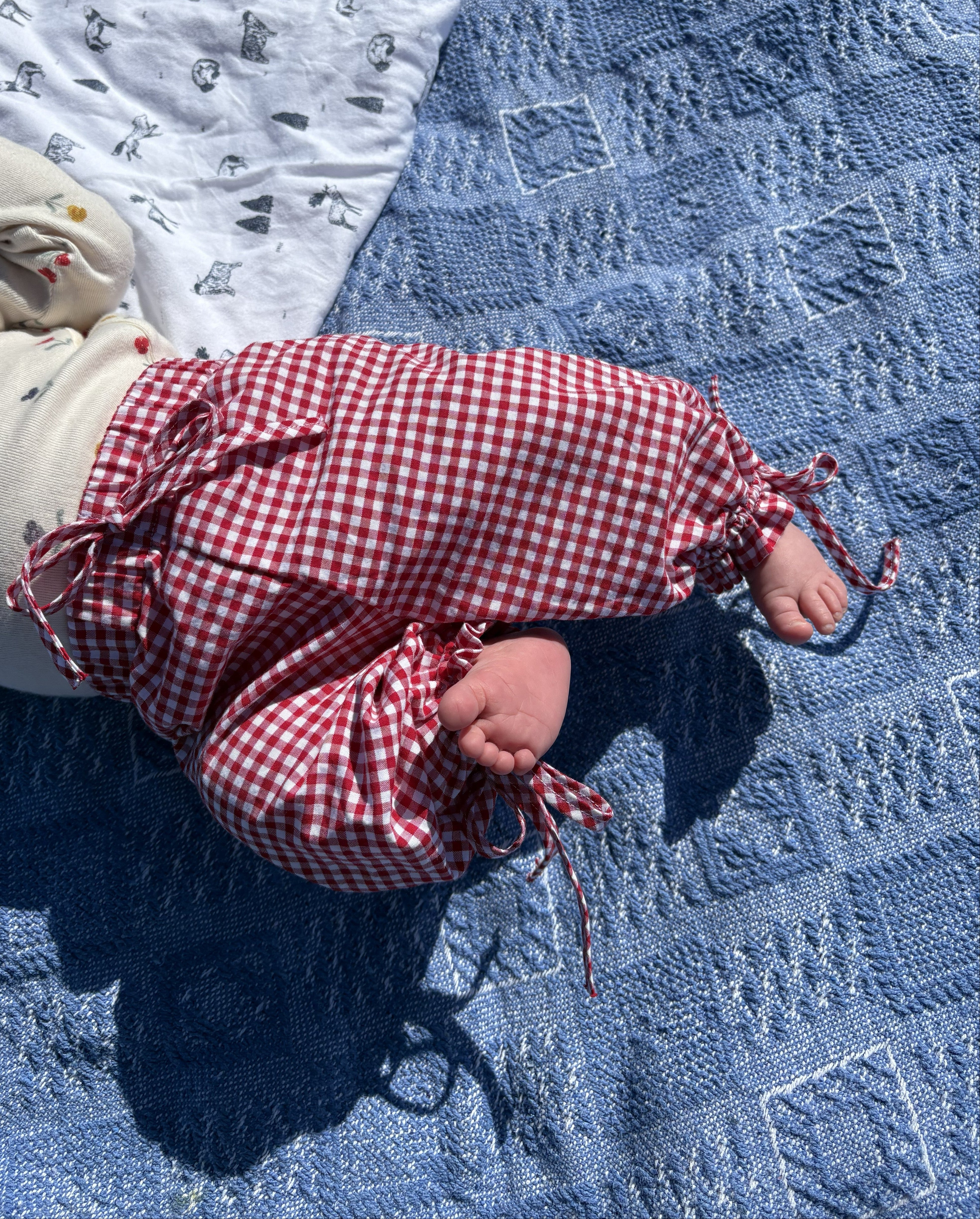 Baby lying on a blue textured blanket wearing a red/white gingham pants.